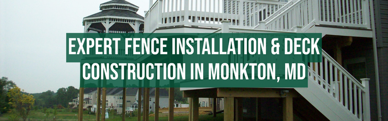 A large green rectangular sign with white text that reads: "EXPERT FENCE INSTALLATION & DECK CONSTRUCTION IN MONKTON, MD" is attached to the side of a white house with a staircase leading to a deck. The house has white railings and a multi-level porch. In the background, there are trees and other houses, with an overcast sky.