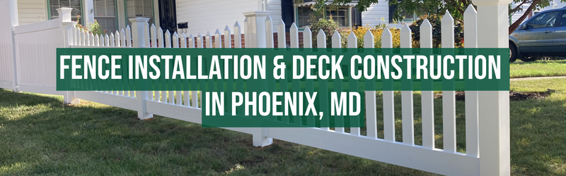A white picket fence runs along the front yard of a house, with a large green banner overlaying the fence that reads: "FENCE INSTALLATION & DECK CONSTRUCTION IN PHOENIX, MD." The background shows a house with a porch, some trees, a driveway with a car, and a well-maintained lawn.