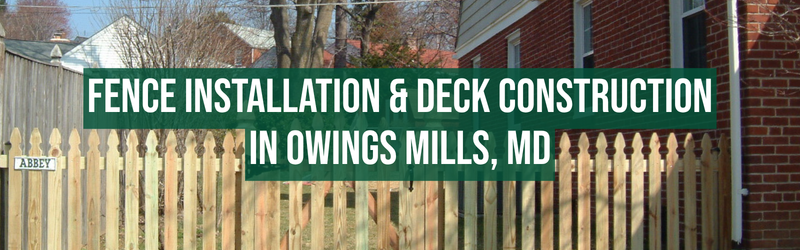 The image shows a wooden fence with vertical pickets in the foreground, and a brick building with white siding in the background. There is a green banner with white text that reads: "FENCE INSTALLATION & DECK CONSTRUCTION IN OWINGS MILLS, MD." To the left, attached to the fence, is a small white sign with black text that says "ABBEY."