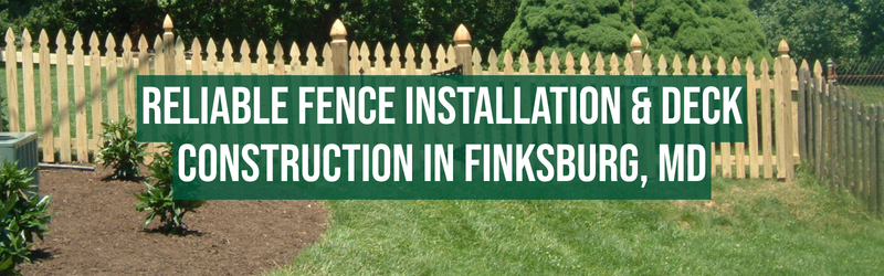 The image shows a wooden picket fence with pointed tops, set in a lush green yard with well-maintained grass and small plants or shrubs along the fence line. Overlaid on the image is a large green banner with white text that reads: "RELIABLE FENCE INSTALLATION & DECK CONSTRUCTION IN FINKSBURG, MD." In the background, there are tall trees and a clear sky, indicating a bright, sunny day.