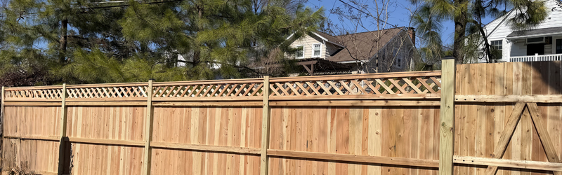 A wooden privacy fence with decorative lattice top runs along a residential backyard, with trees and houses visible in the background under a clear blue sky. A wooden privacy fence with decorative lattice top runs along a residential backyard, with trees and houses visible in the background under a clear blue sky.
