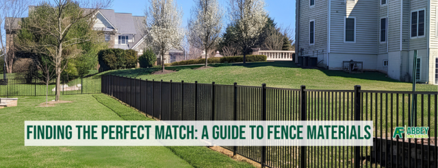 A backyard with a well-maintained lawn, trees in bloom, and a black metal fence separating different sections. There are multi-story houses in the background under a clear blue sky. The image features a banner at the bottom with the text "Finding the Perfect Match: A Guide to Fence Materials" and the Abbey Fence & Deck Co. logo.