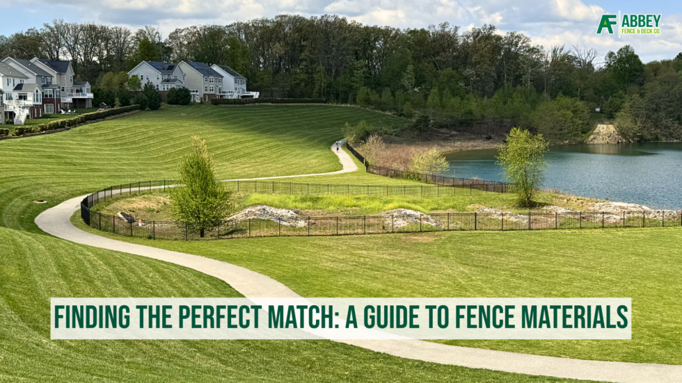 A backyard with a well-maintained lawn, trees in bloom, and a black metal fence separating different sections. There are multi-story houses in the background under a clear blue sky. The image features a banner at the bottom with the text "Finding the Perfect Match: A Guide to Fence Materials" and the Abbey Fence & Deck Co. logo.