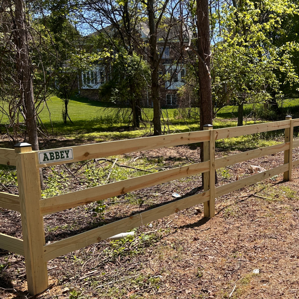 A newly built wooden fence with three horizontal rails and vertical posts, set along a dirt path with some small plants and leaves on the ground. A small sign on the fence reads "ABBEY." Behind the fence, there are trees with green leaves and a house partially visible in the background, surrounded by a lush, grassy yard.