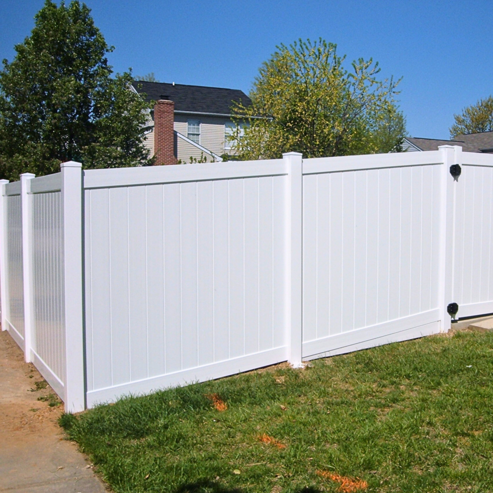 A white vinyl privacy fence with vertical panels and sturdy posts, enclosing a backyard area. The fence is set against a backdrop of green trees and a house with a dark roof, under a bright blue sky. The grass in front of the fence is green and well-maintained.