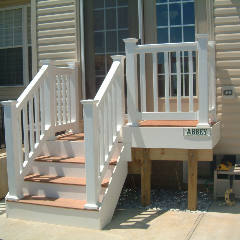 The image shows a small, elevated white porch or deck with stairs leading up to it, attached to a house with beige siding and large windows. The deck features a railing with vertical balusters and a small gate at the top of the stairs. The word "ABBEY" is displayed on a sign attached to the front of the deck. The deck is supported by wooden posts, and there are some gardening tools and supplies visible on the ground nearby.
