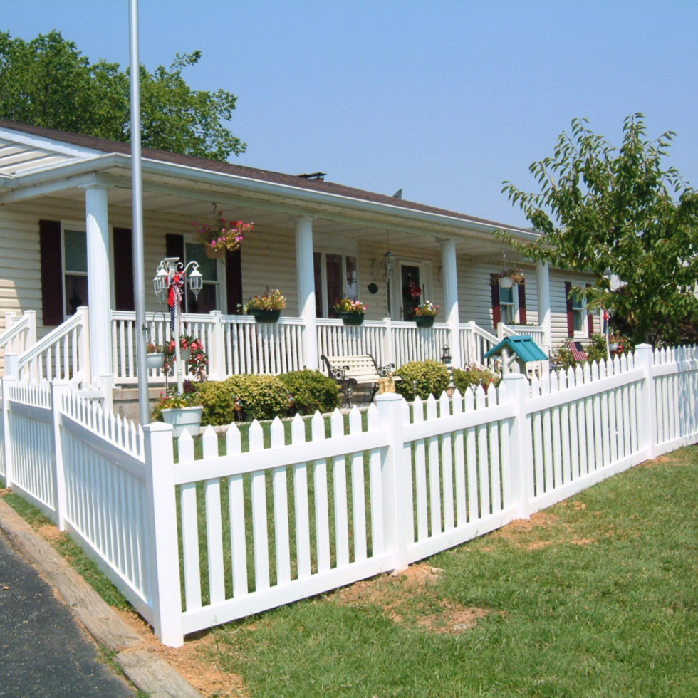 A white picket fence surrounds a well-maintained front yard with green grass. Behind the fence, there is a porch with white columns, flower pots, hanging baskets, and garden decorations. The house has beige siding with dark shutters, and a small tree is visible on the right side of the image. The sky is clear and blue, indicating a sunny day.