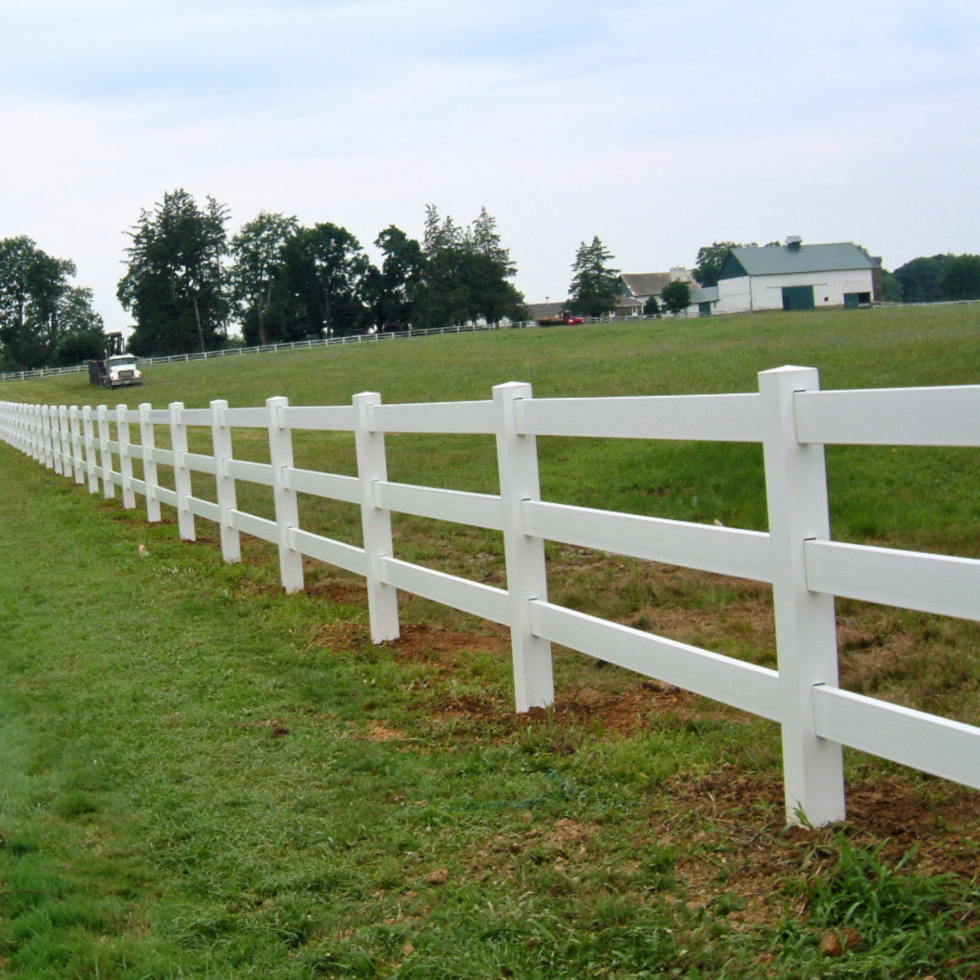 A white PVC paddock fence runs along a grassy field in a rural setting. In the background, there are trees, a few farm buildings, and a golf cart on the grass. The sky is partly cloudy, and the scene appears peaceful and well-maintained.