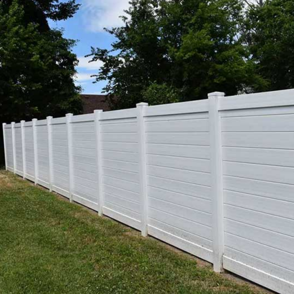 A long, white, privacy fence made of horizontal panels with evenly spaced vertical posts runs along a grassy yard. Behind the fence, tall green trees and a partly cloudy blue sky are visible, creating a peaceful outdoor scene.