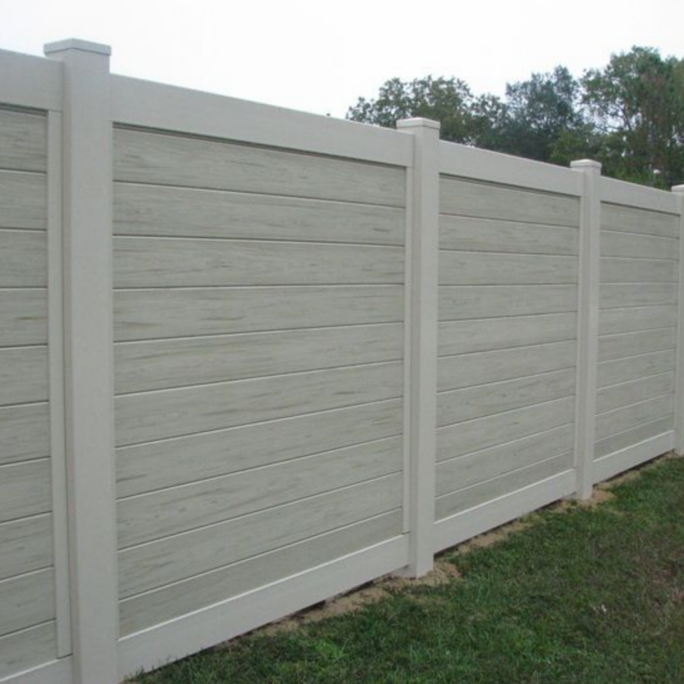 A white privacy fence made of horizontal panels with vertical support posts runs along a grassy area. The fence appears new and well-maintained, with evenly spaced posts and clean, straight panels. In the background, trees and a partly cloudy sky are visible.