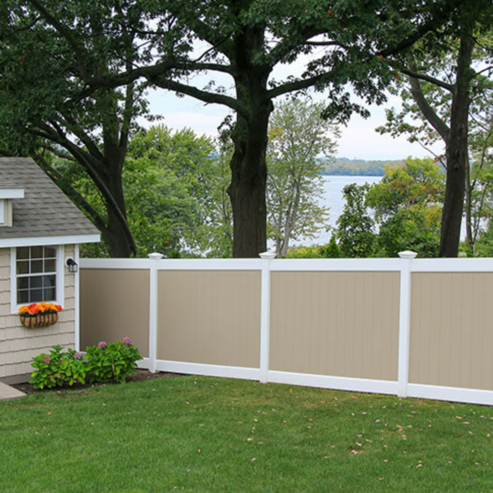 A beige privacy fence with white posts and trim runs along a well-maintained grassy yard. To the left, part of a house with a small window and flower box filled with orange flowers is visible. Behind the fence, there are large trees with lush green foliage, and in the background, a body of water can be seen through the trees. The scene appears peaceful and scenic, suggesting a backyard with a view of a lake or river.