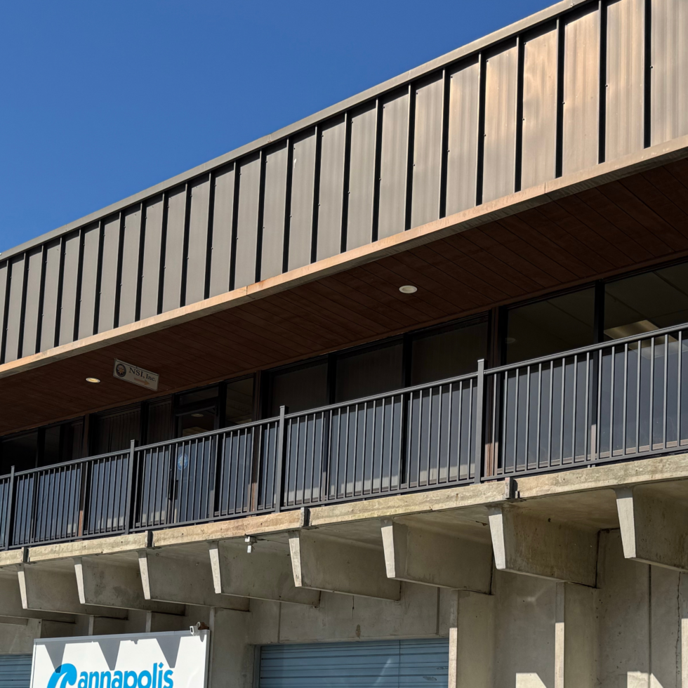A modern building with a concrete base, featuring balconies with black metal railings, and a facade with vertical metal panels. The sky above is clear and blue, and there is a sign at the bottom that reads "Annapolis."