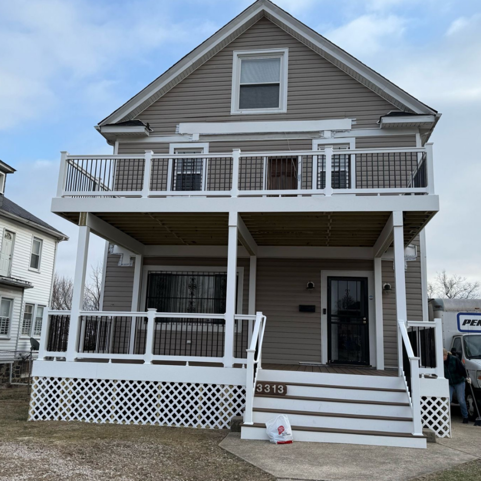 The image shows a two-story house with a beige exterior and white trim. It features a front porch with stairs leading up to the main entrance, which has a black door. The house has a large balcony on the second floor with white railings, supported by wooden posts. The house number "3313" is displayed on the front steps. There is a bag placed at the bottom of the stairs, and a person standing near a van on the right side of the image. The sky is partly cloudy.