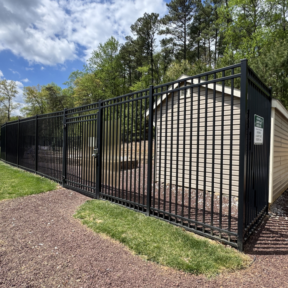 A black metal fence borders a landscaped area with mulch and small plants, adjacent to a grassy lawn. In the background, there are tall trees with green foliage and a partly cloudy sky overhead. The scene appears to be outdoors on a bright, sunny day.