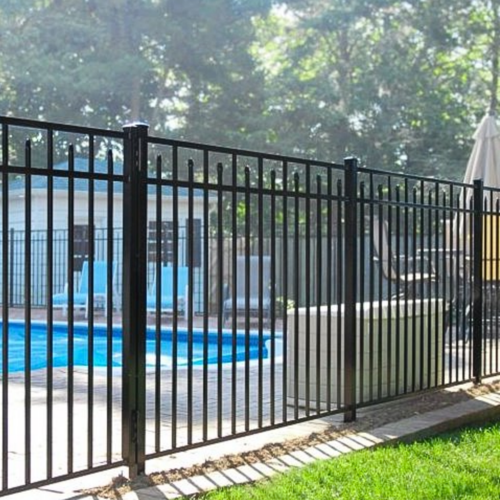 A black metal fence with vertical bars surrounds a backyard pool area. Behind the fence, there are blue lounge chairs and a pool with clear blue water. In the background, there are trees with green foliage, a white umbrella, and outdoor furniture, creating a cozy outdoor setting on a sunny day.