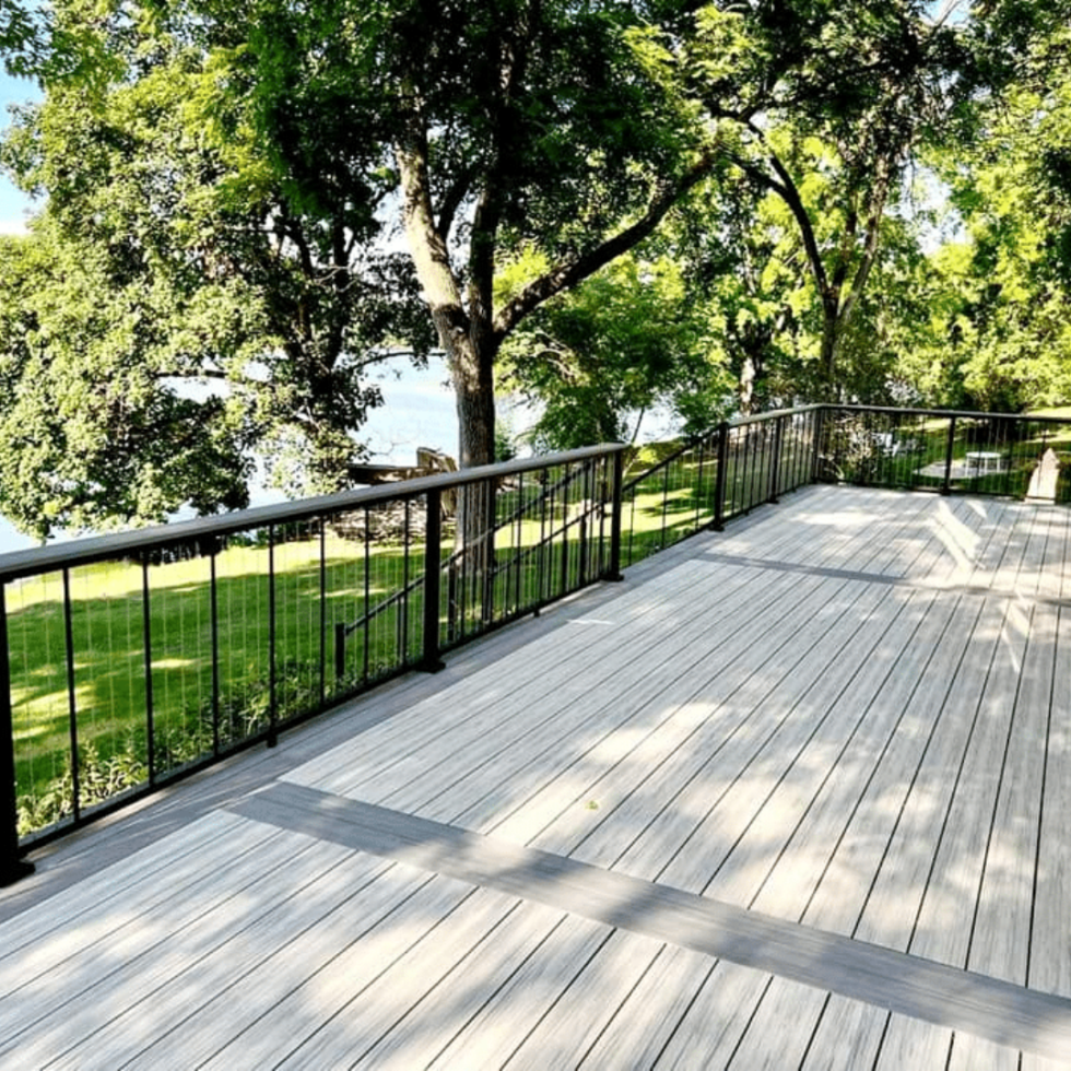 The image shows a spacious outdoor deck with light-colored wooden flooring and black metal railing. The deck extends outward and is surrounded by lush green trees, providing a shaded and serene environment. In the background, there is a grassy area with more trees and a glimpse of a body of water through the foliage. The scene captures a peaceful, natural setting on a bright, sunny day.