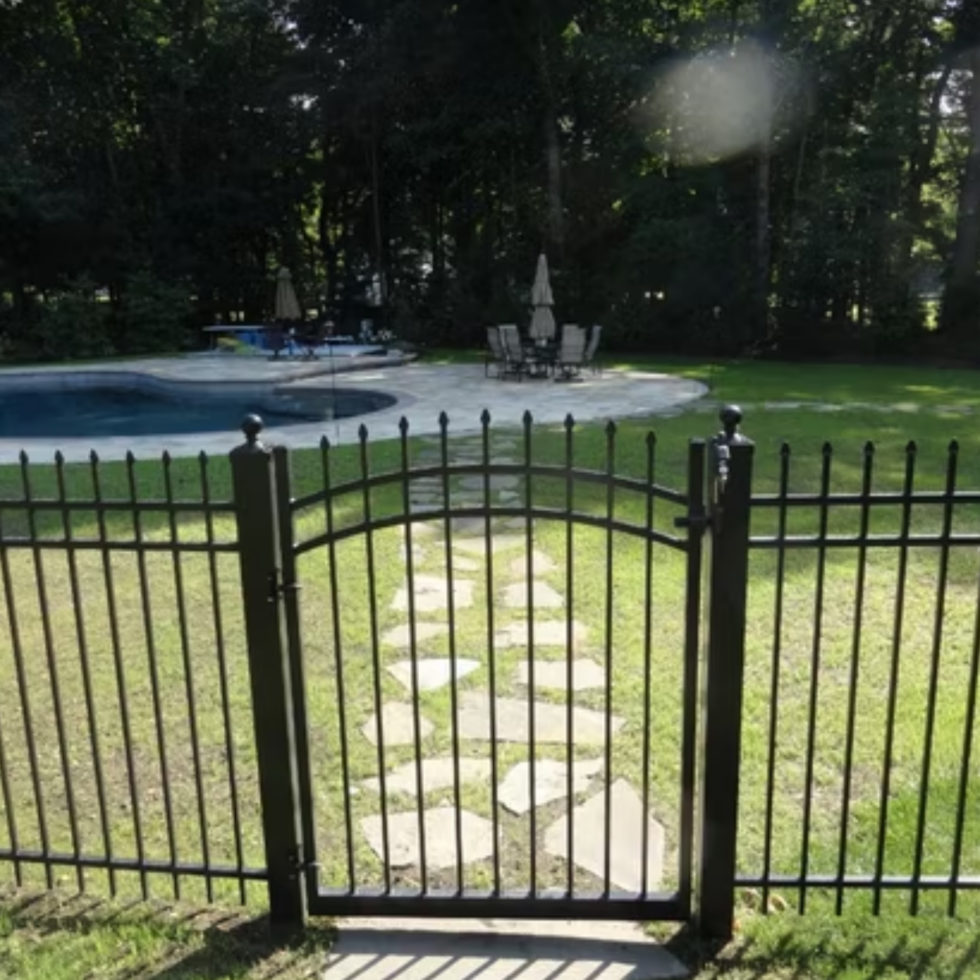 A black metal fence and gate with decorative finials encloses a backyard with a grassy lawn. Behind the gate, there is a stone pathway leading to a patio area with a round table, chairs, and a large umbrella. In the background, there is a swimming pool and a wooded area with tall trees providing shade. The scene appears to be a peaceful, private outdoor space.
