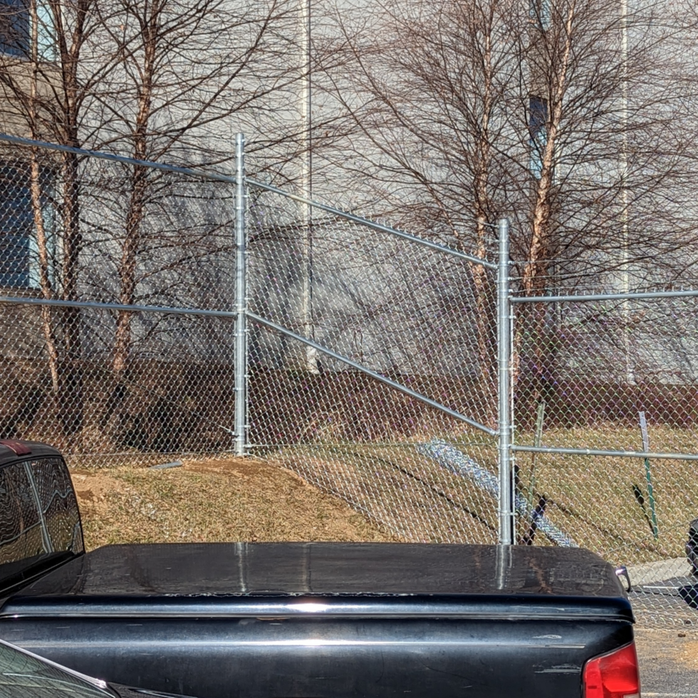 The image shows the rear windshield and trunk of a black car parked behind a chain-link fence. The background features leafless trees and a building with large windows. The sunlight casts shadows on the scene, indicating it is a clear day.