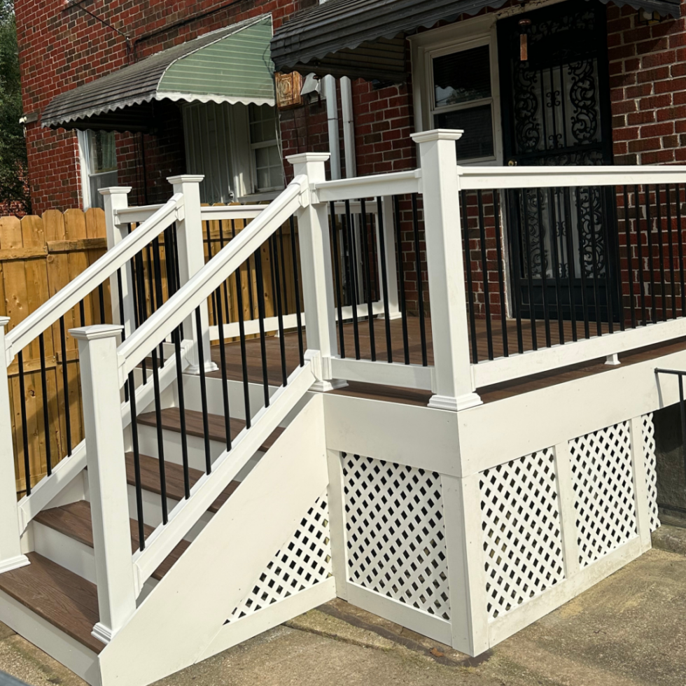 The image shows a newly built outdoor deck attached to a brick house. The deck features white railings with black balusters, a staircase with matching white posts, and a lattice skirting underneath. The deck is elevated, with stairs leading down to a concrete patio. The house has a window with a green and white striped awning, and a black security door with intricate metalwork. There is a wooden fence in the background separating the yard from neighboring properties.