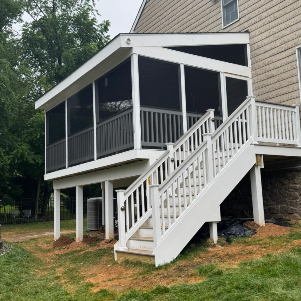 The image shows a screened-in porch or sunroom attached to the back of a house. The structure is elevated on white support posts, with a staircase leading up to a white railing around the porch. The porch is enclosed with black screened panels, and the house has beige siding with a small window visible on the upper level. The area beneath the porch has some exposed dirt and construction materials. Trees and a fence are visible in the background.