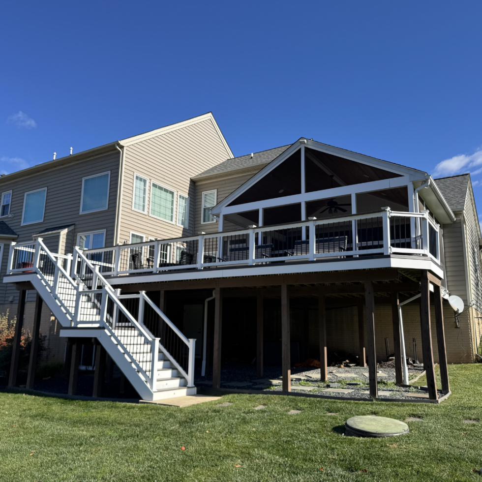 The image shows the back of a large, multi-story house with beige siding and multiple windows. There is a spacious, elevated deck with white railings and stairs leading down to a grassy yard. The deck has a screened-in porch area with a sloped roof, supported by white posts. The area underneath the deck is open and appears to be under construction or storage, with some construction materials visible. The sky above is blue with a few clouds.