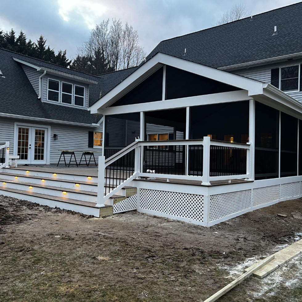 The image shows a modern, multi-level house with a large, covered outdoor deck. The deck features white railings with black balusters, stairs leading down to the yard, and built-in lighting along the stairs. The house has gray siding, multiple windows, and a steeply pitched roof. The yard appears to be under preparation or landscaping, with bare soil and some wooden framing. The overall scene is set during the daytime with a cloudy sky overhead.