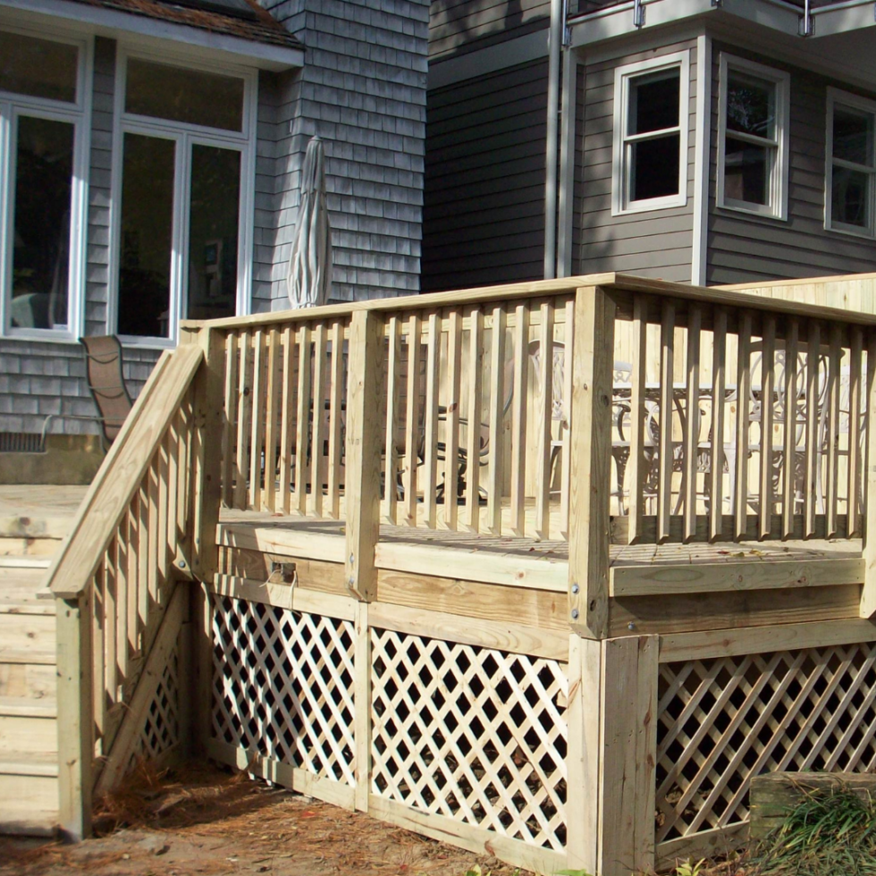 The image shows a newly built wooden deck with a railing, attached to a house. The deck has stairs leading down to the ground, and the underside features lattice panels. The house's exterior has gray siding, and there are large windows and a sliding glass door adjacent to the deck. The area appears to be in the process of construction or renovation.