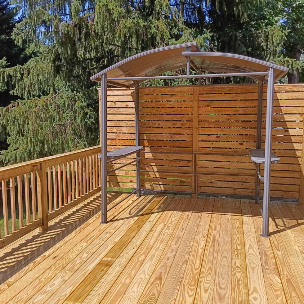 The image shows a wooden deck with a metal and wood structure that appears to be a small outdoor shelter or stand, possibly for a grill or similar item. The deck is enclosed by a wooden railing, and there is a wooden privacy fence behind the structure. In the background, tall green trees provide a natural setting. The sky is clear and sunny.