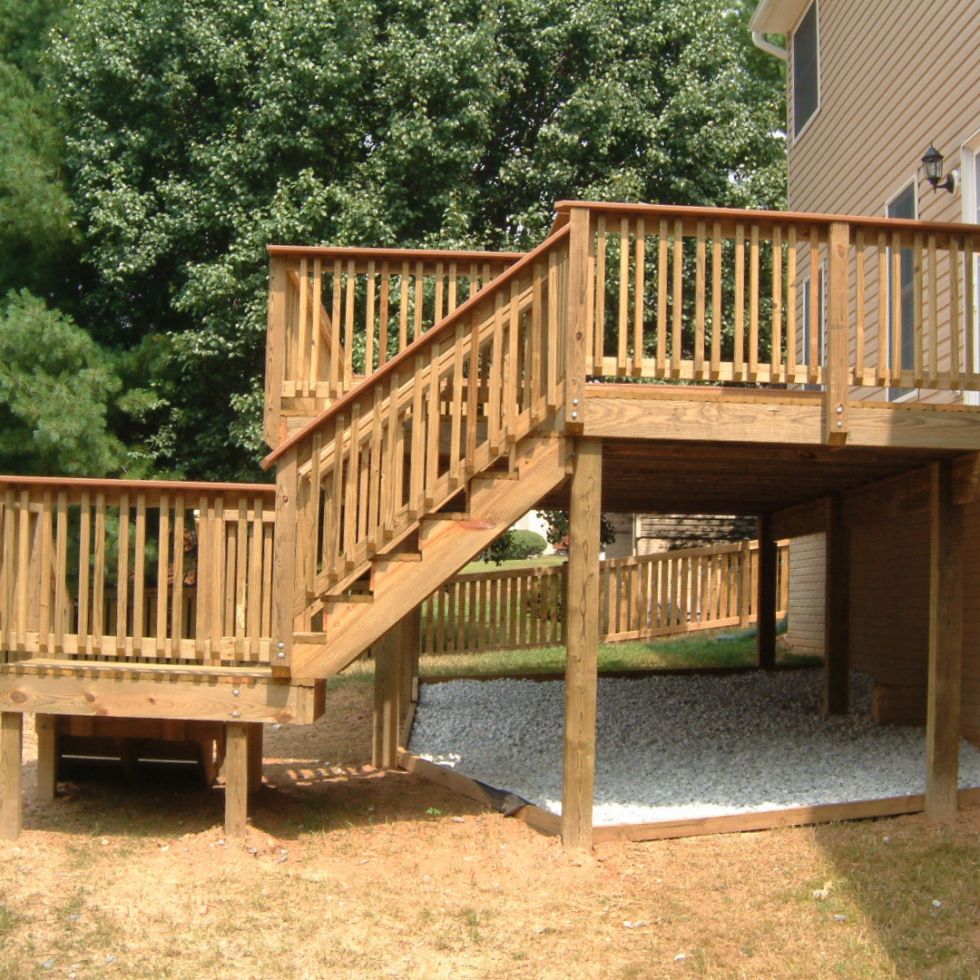 The image shows a newly constructed wooden deck with stairs attached to a house. The deck has railings for safety, and the stairs lead down to the yard. The area beneath the deck is supported by wooden posts, and there is a section filled with white gravel. The background features lush green trees and part of the house's exterior.
