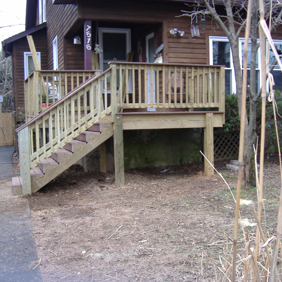 The image shows a newly built wooden deck attached to a house, with a set of stairs leading down to the ground. The deck has railings for safety, and the area underneath the deck is open with support posts. To the right, there is a tree with some stakes and ties around its trunk, likely for support. The yard in front of the deck appears to be in the process of landscaping, with bare soil and some scattered debris. The house has brown siding, and there are windows and a door leading onto the deck.