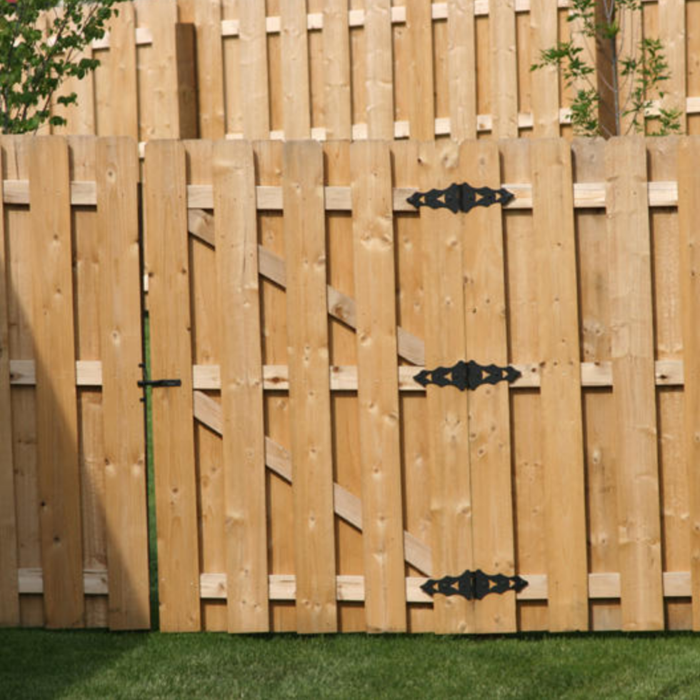 A newly installed wooden privacy fence with vertical planks and decorative black hinges, enclosing a yard with green grass. The fence is made of light-colored wood, with a gate in the center. Some small plants are visible behind the fence, and the scene is brightly lit, suggesting daytime.