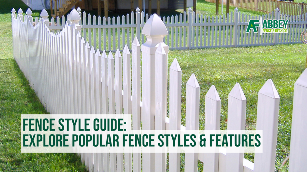 The image features a white picket fence with pointed tops, running along a lush green lawn. The fence has decorative posts with rounded caps, and a small gate is visible in the foreground. In the background, there is a larger, light blue fence enclosing a yard with trees and a wooden deck. The top right corner displays the Abbey Fence & Deck Co. logo, and the bottom left corner contains text that reads: "FENCE STYLE GUIDE: EXPLORE POPULAR FENCE STYLES & FEATURES."