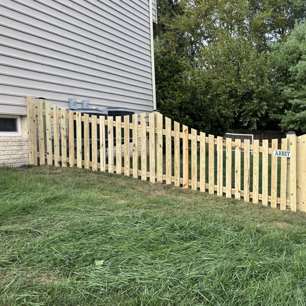 A newly constructed wooden fence with vertical slats, installed along the side of a house with beige siding and brick foundation. The fence features a small gate labeled "ABBEY" and is situated on a grassy lawn. Behind the fence, there are trees and a neighboring yard, with some HVAC units visible near the house. The scene appears to be in a suburban residential area.