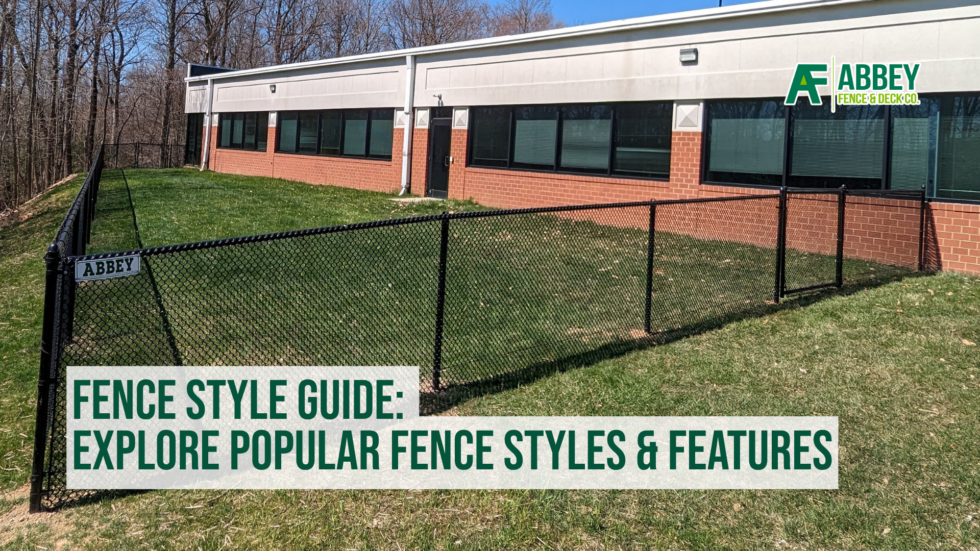 A black chain-link fence with a small sign that says "ABBEY" in a grassy area next to a building with large windows and a brick base. The building has a white exterior with a logo in the top right corner that reads "ABBEY FENCE & DECK CO." In the foreground, there is a text overlay that says, "FENCE STYLE GUIDE: EXPLORE POPULAR FENCE STYLES & FEATURES."