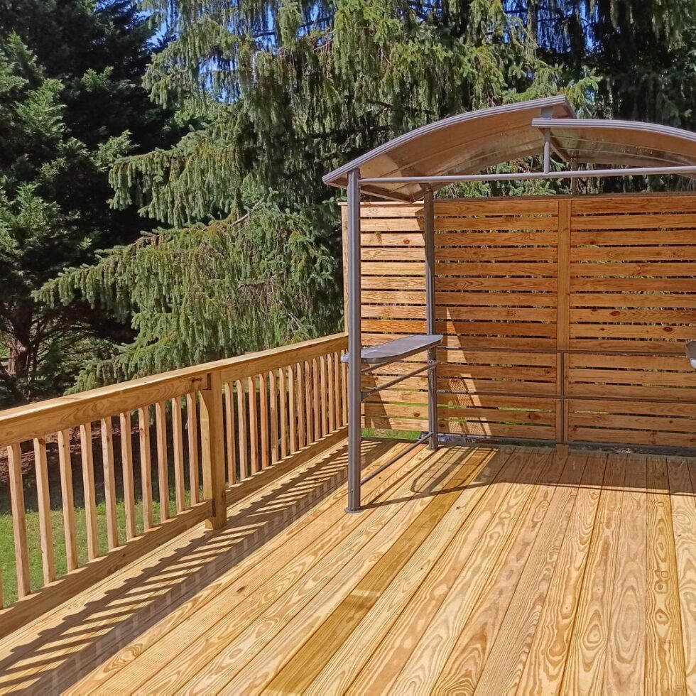 Spacious pressure-treated wood deck featuring a privacy screen, covered grill station, and safety railings in a backyard surrounded by mature trees.