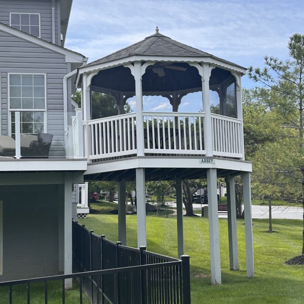 Custom elevated screened gazebo deck with white railings and a shingled roof, attached to a residential home and overlooking a landscaped backyard.