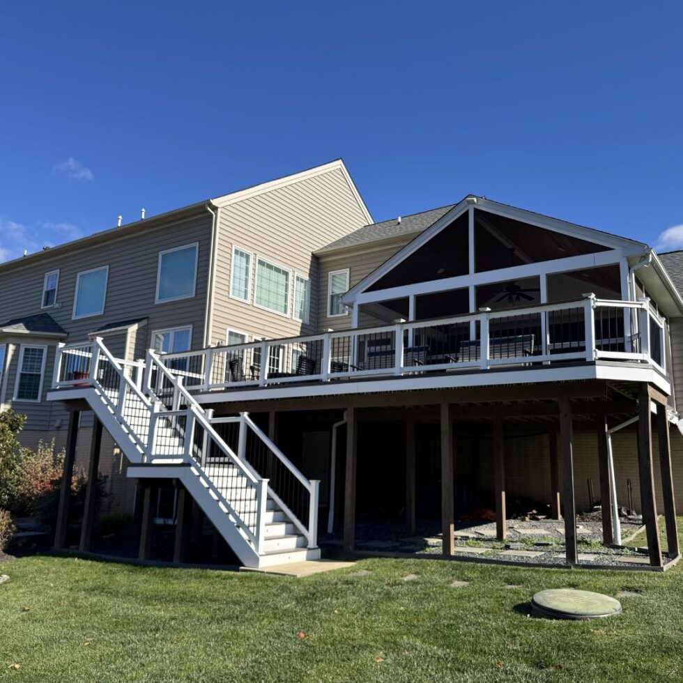 Large elevated backyard deck with a covered porch, white aluminum railings, staircase access, and open under-deck space attached to a multi-story home.